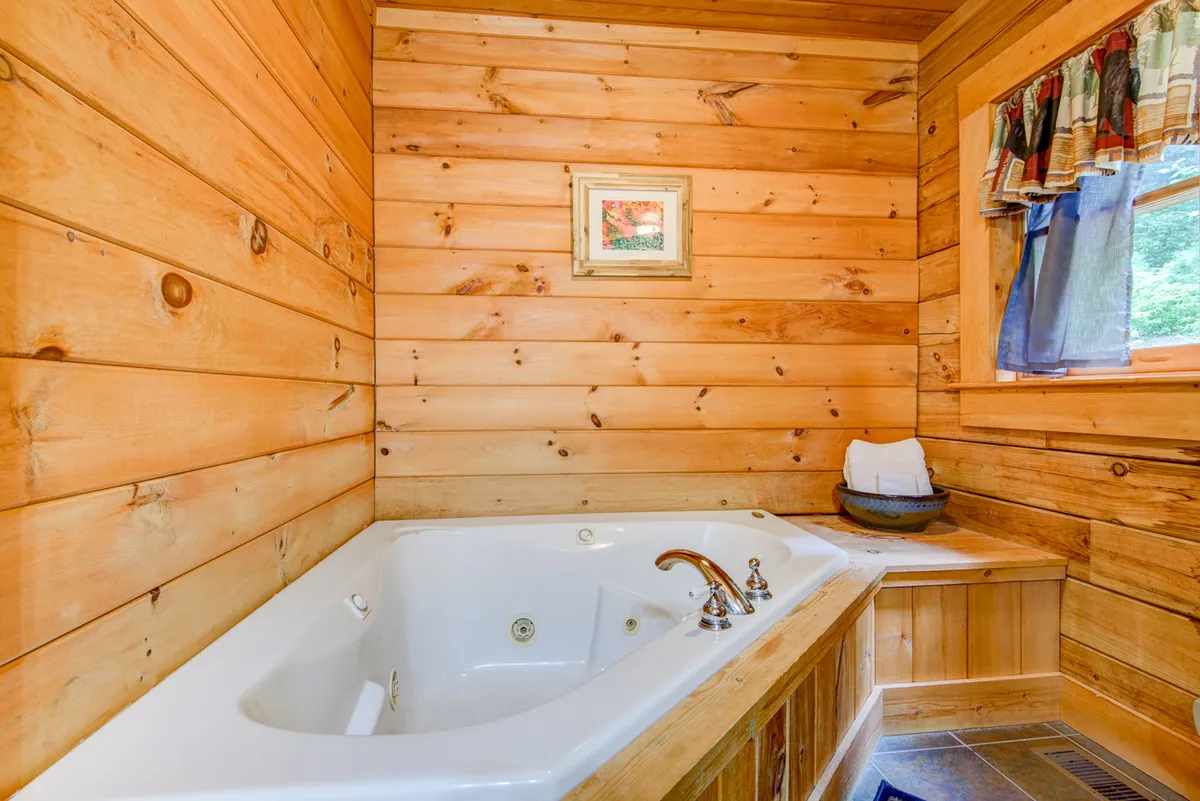 A tranquil bathroom with a jacuzzi tub set against wooden walls, accented by framed artwork and a window with curtains. The space exudes spa-like serenity, perfect for unwinding after adventure-filled days.