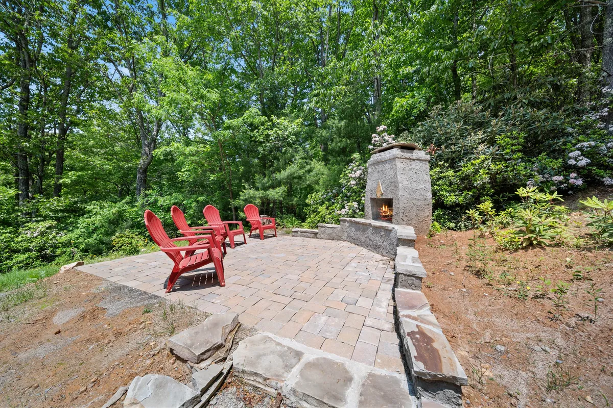 A cozy outdoor space with red Adirondack chairs arranged around a striking stone fireplace on a paved patio. Lush greenery creates a sense of seclusion, making it an ideal spot for evening conversations or fireside gatherings under the stars.