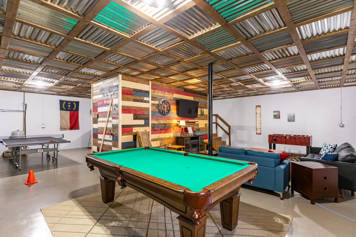 A vibrant recreation room featuring a pool table under modern lighting, a colorful accent wall, a comfortable seating area with plush chairs, and a large television mounted on the wall. The space invites social gatherings, with open sightlines and energetic décor.