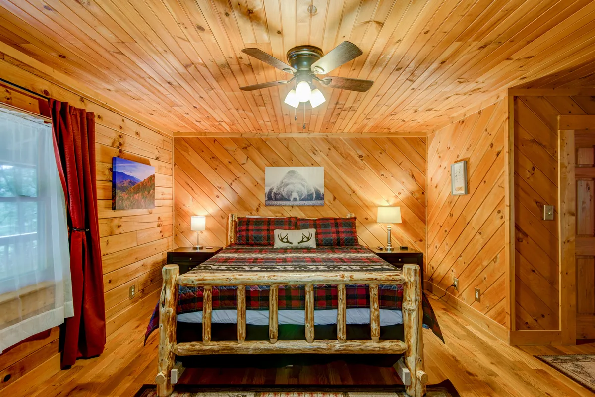 A cozy bedroom with a log-framed bed dressed in plaid bedding, warm wooden wall paneling, rustic artwork, and a ceiling fan. Soft light spills in from a nearby window, enhancing the inviting atmosphere.