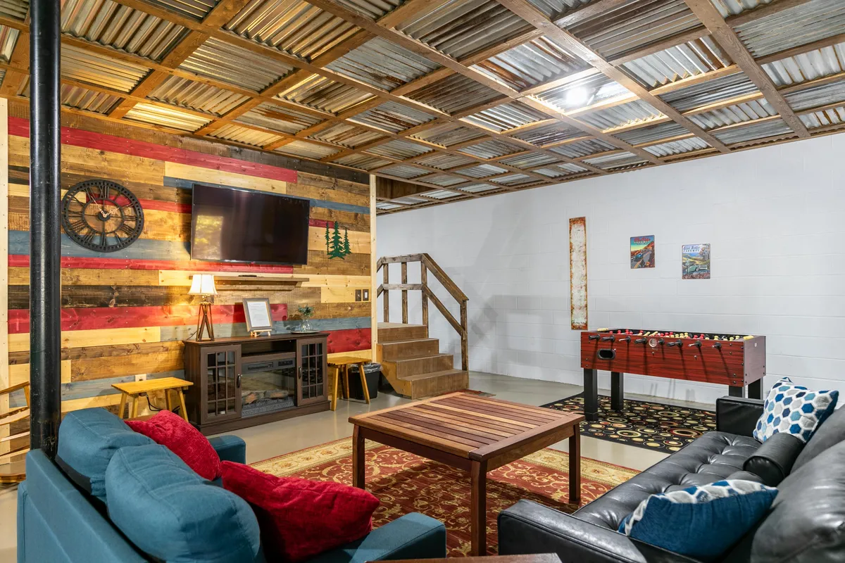 A lively corner of the recreation room with a foosball table, large TV, eclectic wooden wall with colorful planks, a cozy seating area, and a staircase leading to another level. The design balances rustic charm with modern comfort, ideal for socializing and playing games.