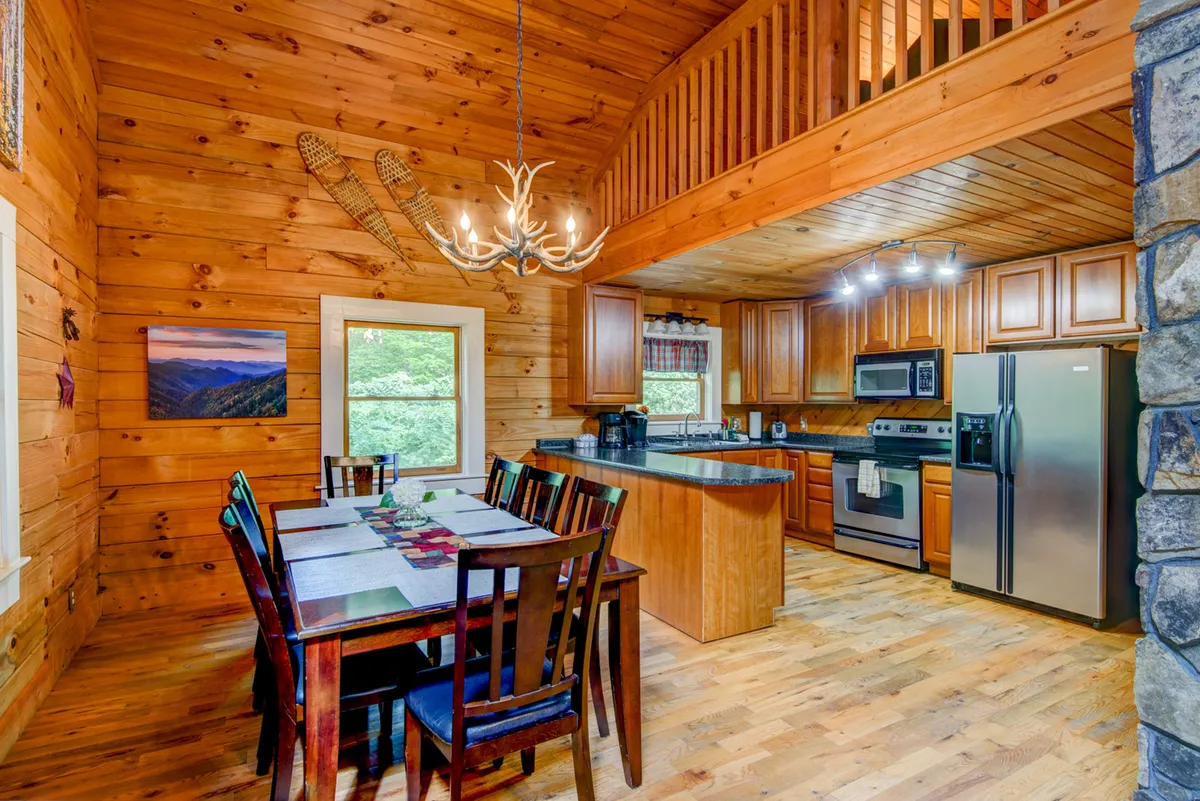 A bright, wood-paneled dining area connected to a spacious kitchen, with a large table under a classic chandelier. The kitchen island and surrounding cabinetry are visible, inviting guests to cook and share meals.
