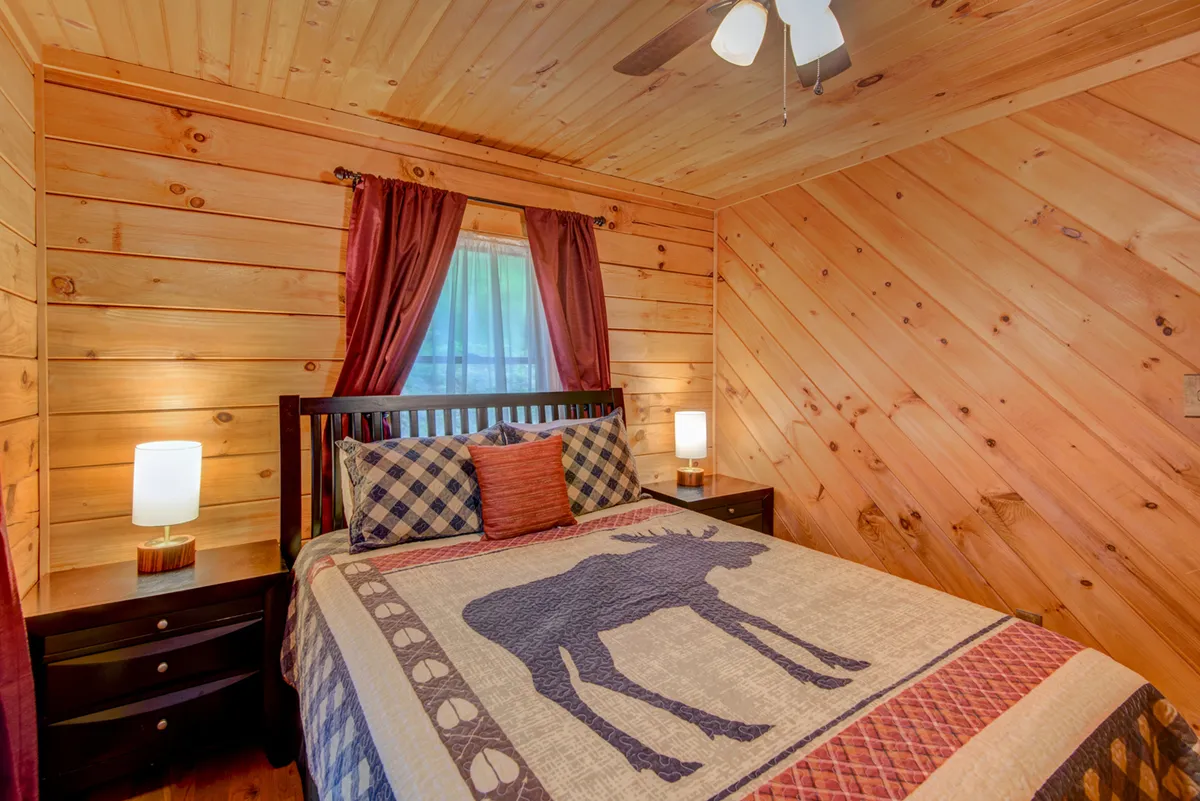 A rustic bedroom showcasing a bed covered with a moose-patterned blanket, wooden paneled walls, and curtains framing a sunlit window. The setting is intimate, inviting, and ideal for quiet rest or reading.