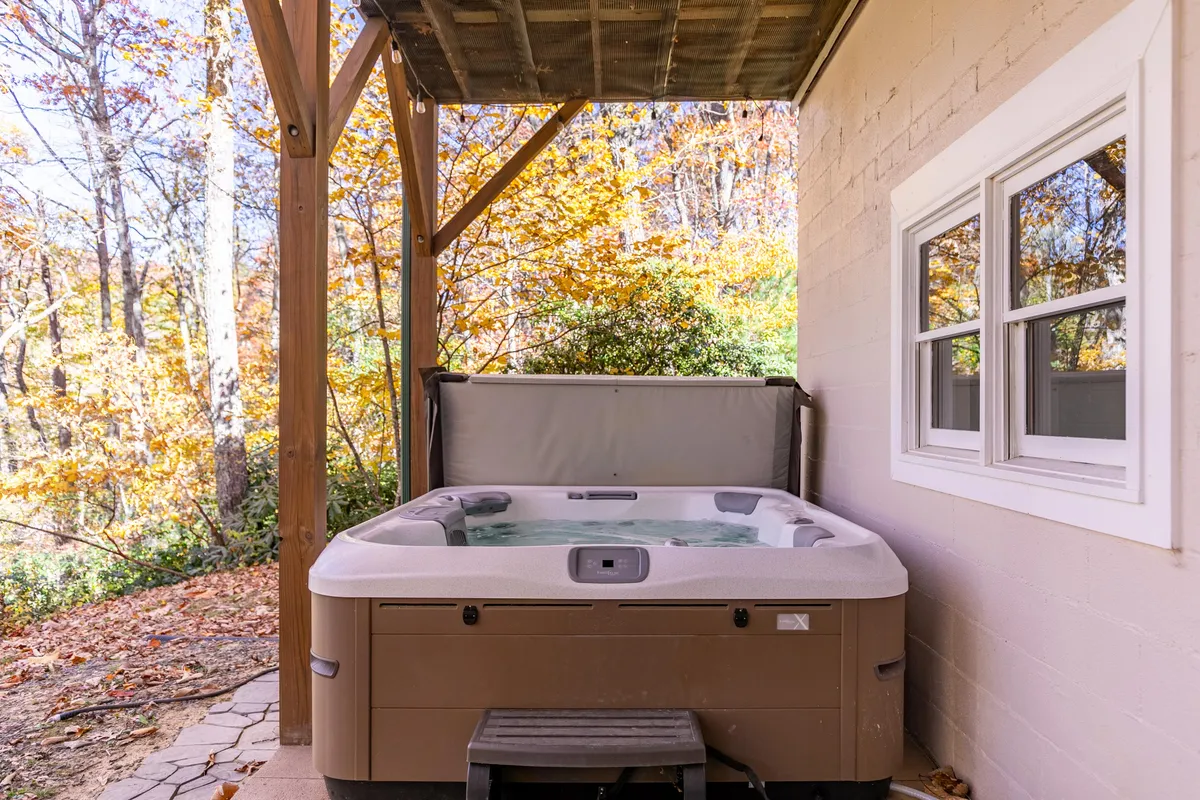 A serene exterior scene featuring a bubbling hot tub nestled against the cabin, surrounded by mature trees and greenery. A stone pathway leads to the shaded area underneath the roof, inviting guests to relax in total privacy while soaking up the natural beauty.