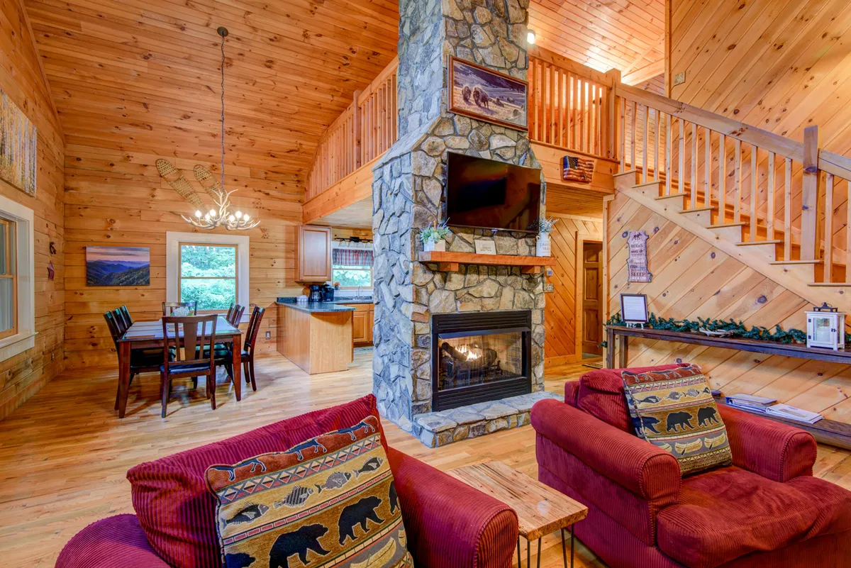 A welcoming interior scene highlighting the seamless transition between the living area and dining space. The stone fireplace and wooden staircase anchor the space, while a dining table with red chairs promises convivial meals for a group.