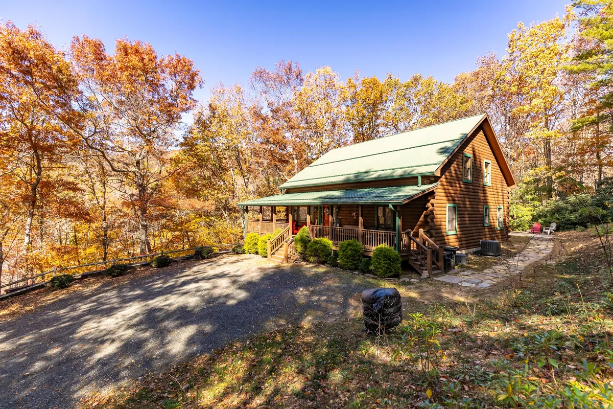 The image shows the log cabin surrounded by vibrant autumn foliage, with the front porch featuring inviting seating. A gravel driveway winds up to the cabin, emphasizing both the natural beauty of the changing seasons and the secluded, adventurous approach to the property.