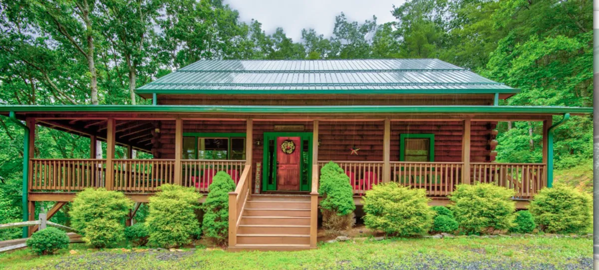 The image showcases a cozy log cabin with a green metal roof, nestled among tall trees on a landscaped plot. The front porch, adorned with a decorative wreath and surrounded by lush shrubs, invites guests to relax and soak in the peaceful woodland setting. Warm lighting and rustic touches evoke a sense of welcome and tranquility.