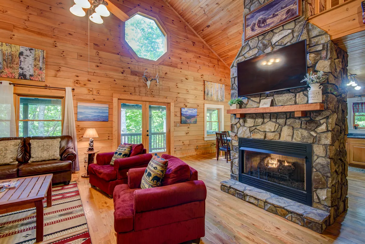 A warm, inviting great room featuring a dramatic stone fireplace flanked by large windows that let in abundant natural light. Red armchairs are arranged for cozy gatherings, and the room's wooden ceiling and octagonal window add rustic character.
