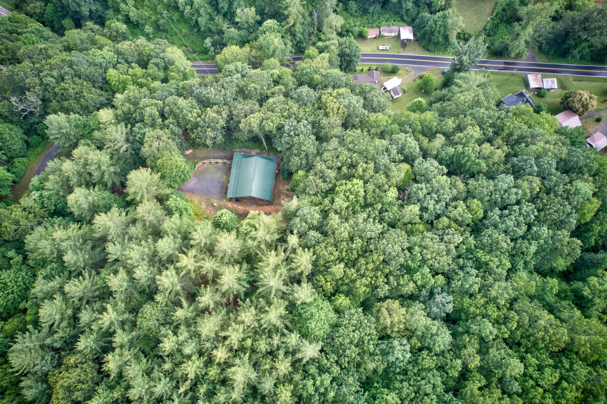An aerial view of a cabin nestled within dense forest greenery, with a surrounding road hinting at the vastness and privacy of the 15-acre property. The scene is peaceful, inviting exploration and outdoor adventure.