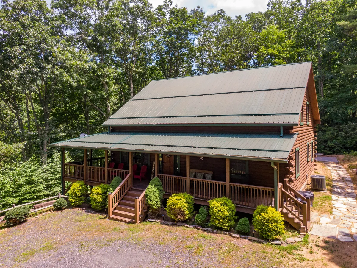 The log cabin structure with a green metal roof, front porch seating, and a landscaped garden. The scene exudes warmth and welcome, with the porch offering a spot for morning coffee or evening relaxation while surrounded by nature.